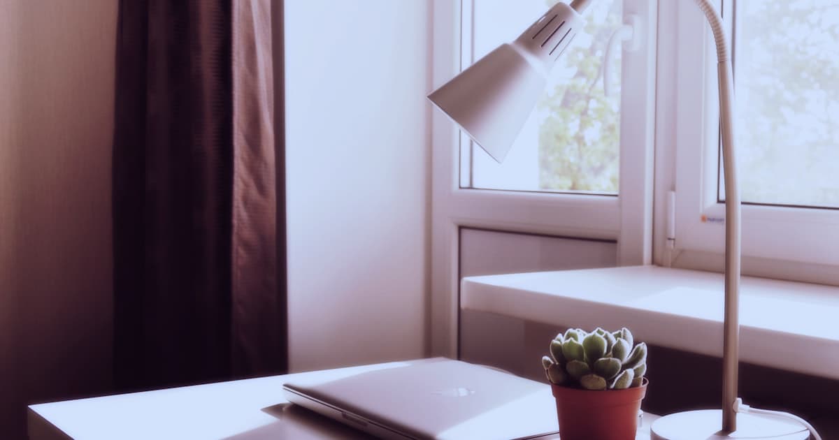 A minimalist home office desk with a desk lamp, open laptop, and small plant in soft natural light